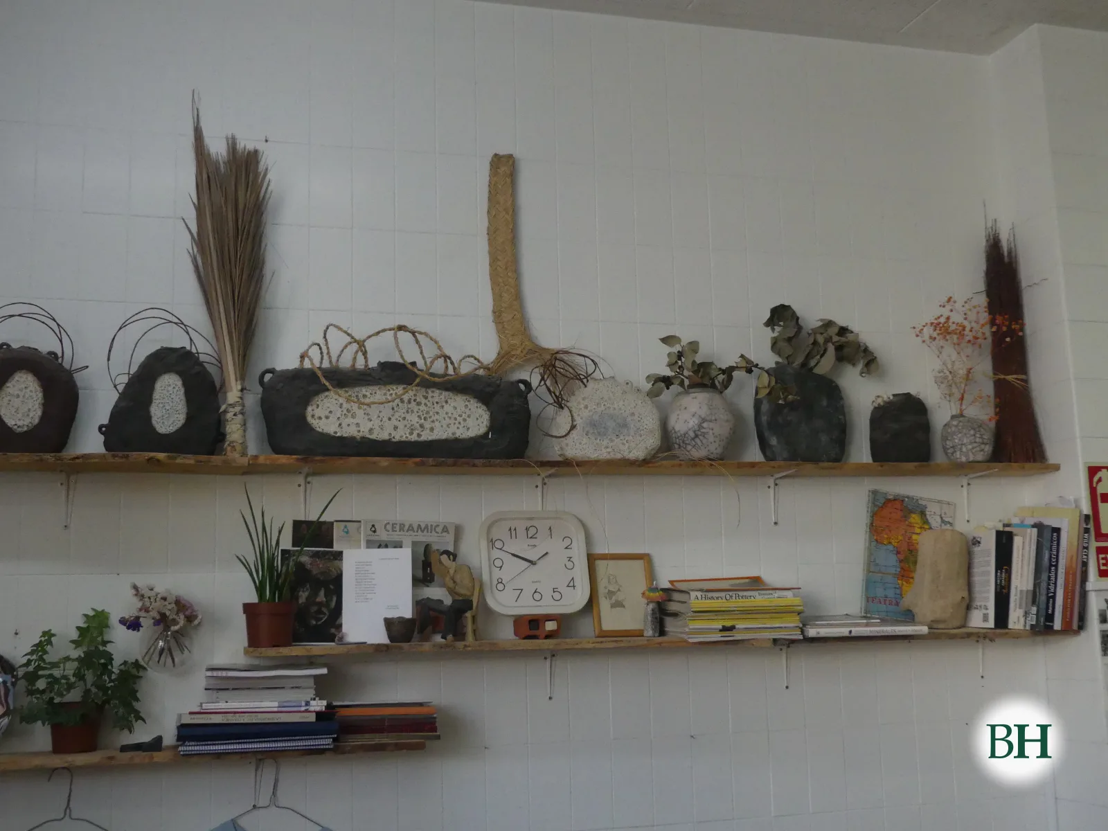 Estante con una fila de piezas de cerámica oscuras y blancas con mimbre, esparto y ramas secas encima, sobre otro estante con libros, plantas y un reloj.