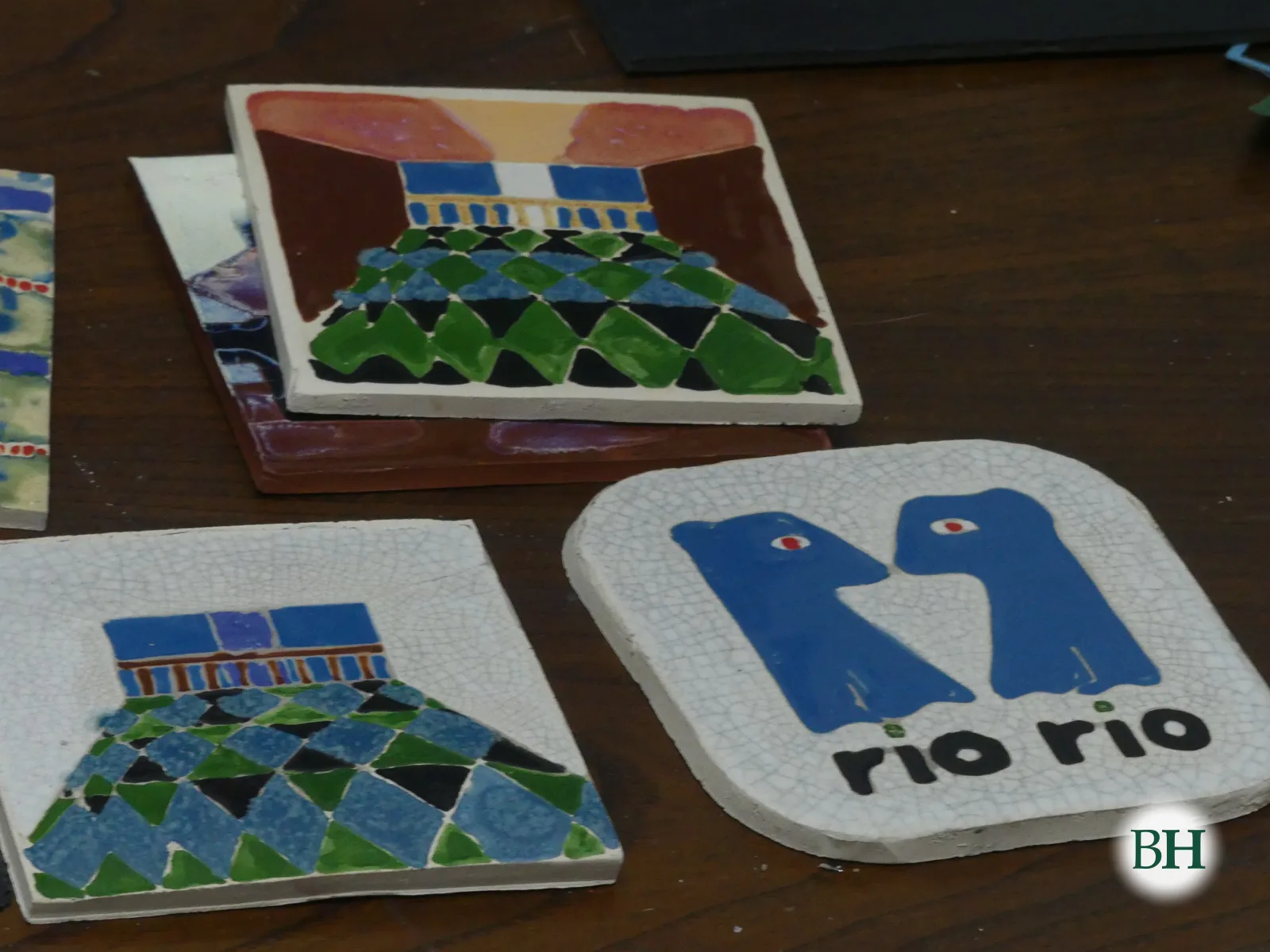 Cuatro azulejos esmaltados sobre una mesa de madera: dos con rombos verdes, azules y negros bajo un balcón azul, y uno cuadrado blanco con el logotipo azul de Río Río, dos figuras enfrentadas.