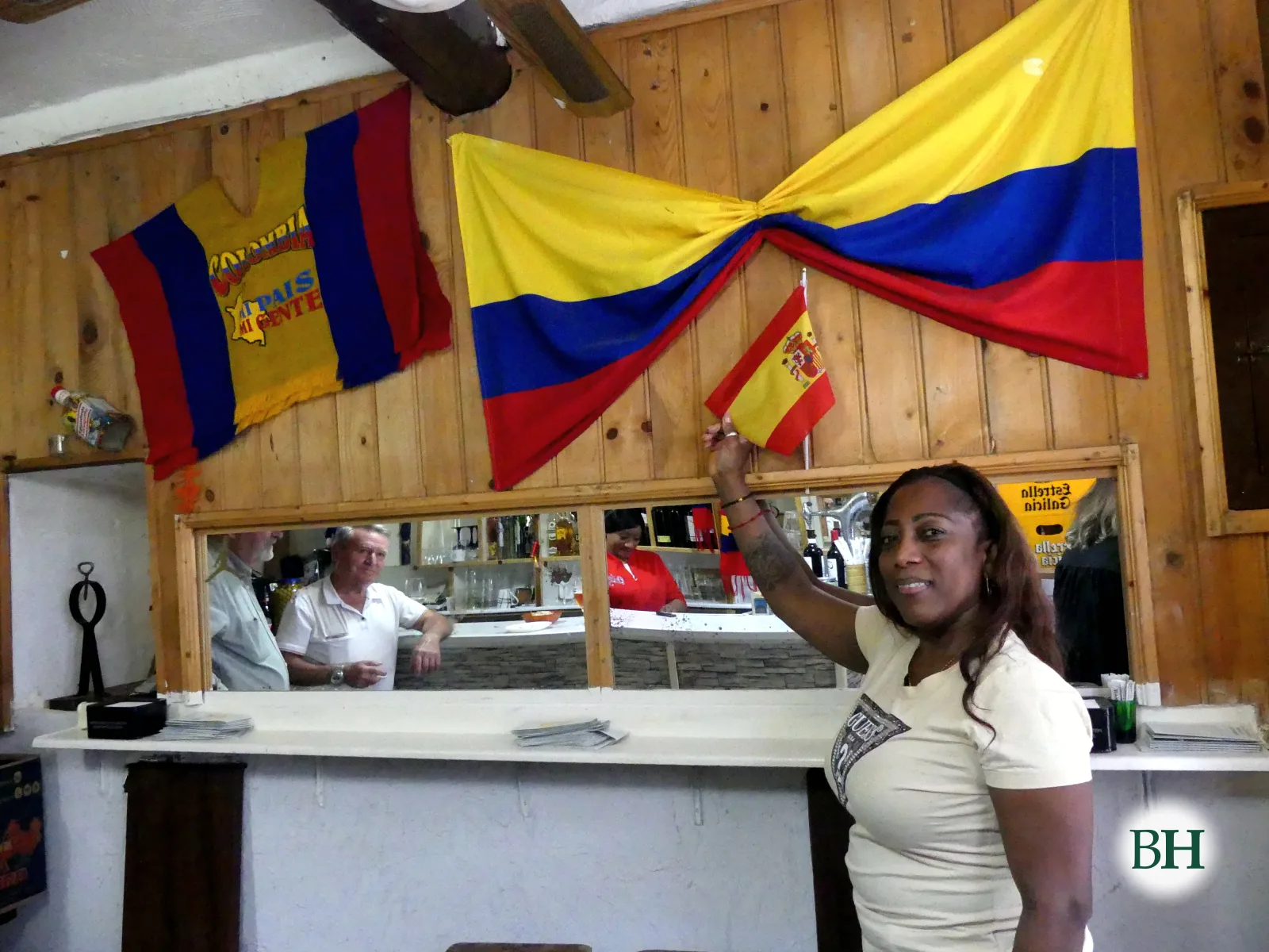 Maricela Palacio Cabeza sorrí e ergue unha bandeira de España pequena diante de dúas bandeiras grandes de Colombia colgadas na parede de madeira do bar. Ao fondo, a barra con clientes.