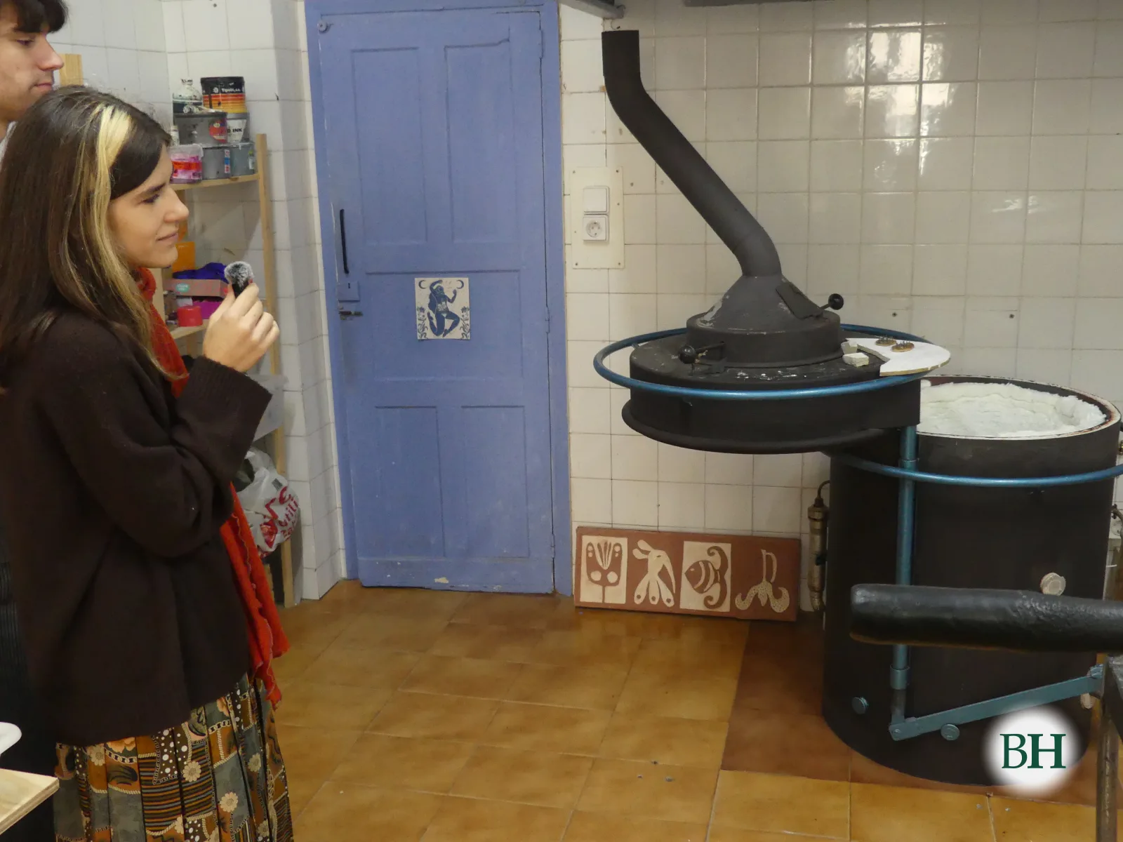 Andrea Aguafría, con un micrófono en la mano, delante de un horno de cerámica de gas, cilíndrico y negro, con la tapa abierta y una chimenea que sube hacia la campana.