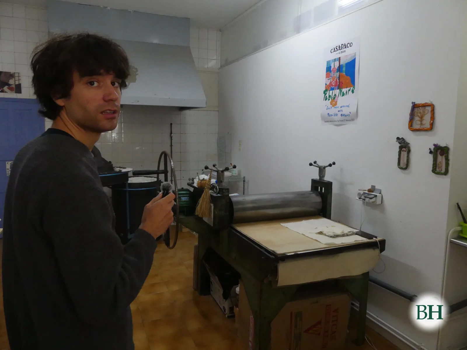Pablo Naire, con un micrófono en la mano, al lado de un tórculo verde con el cilindro metálico y la pletina cubierta de papel. En la pared, el cartel de una exposición suya en Casa Paco.