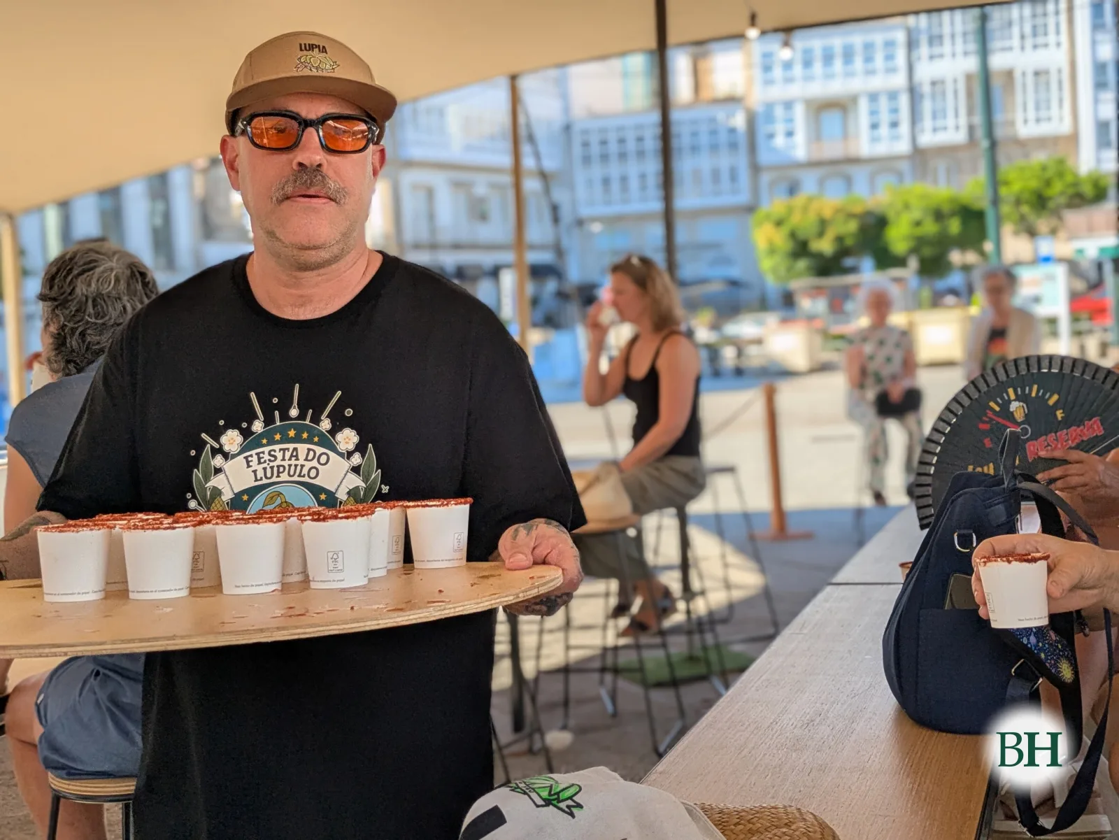 Un hombre con la camiseta de la Fiesta del Lúpulo lleva una bandeja con vasos de degustación bajo la carpa