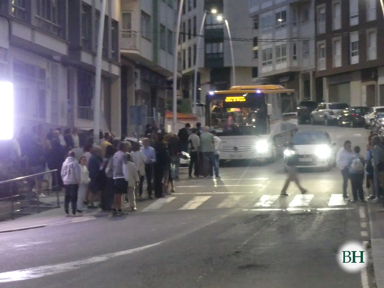 Grupo de persoas cruzando un paso de peóns en Miño ao amencer, co autobús detido na parada detrás.