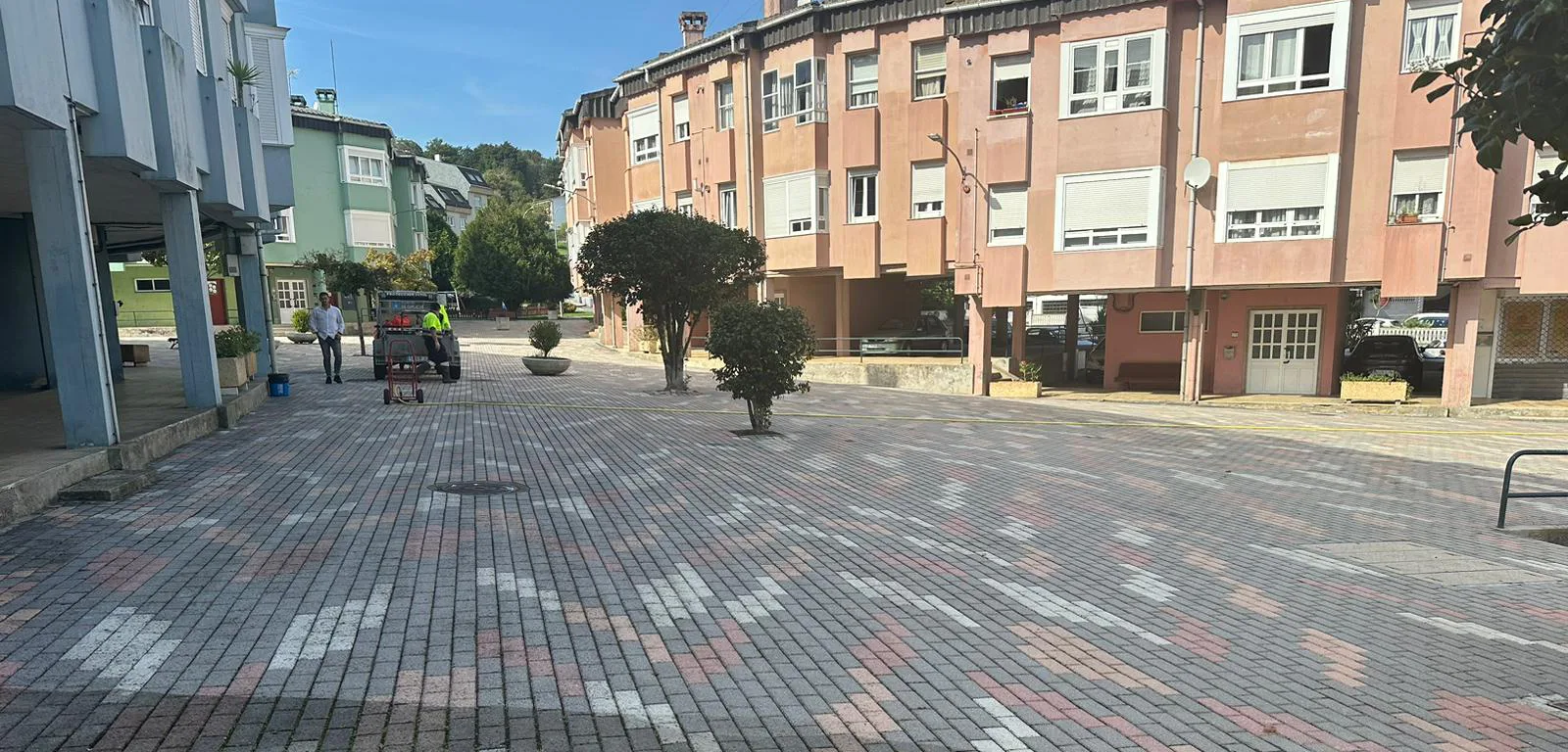 Zona central da urbanización da Condesa: unha ampla superficie pavimentada con adoquín de cores entre edificios de vivendas de catro alturas, con dúas árbores e algunhas xardineiras.