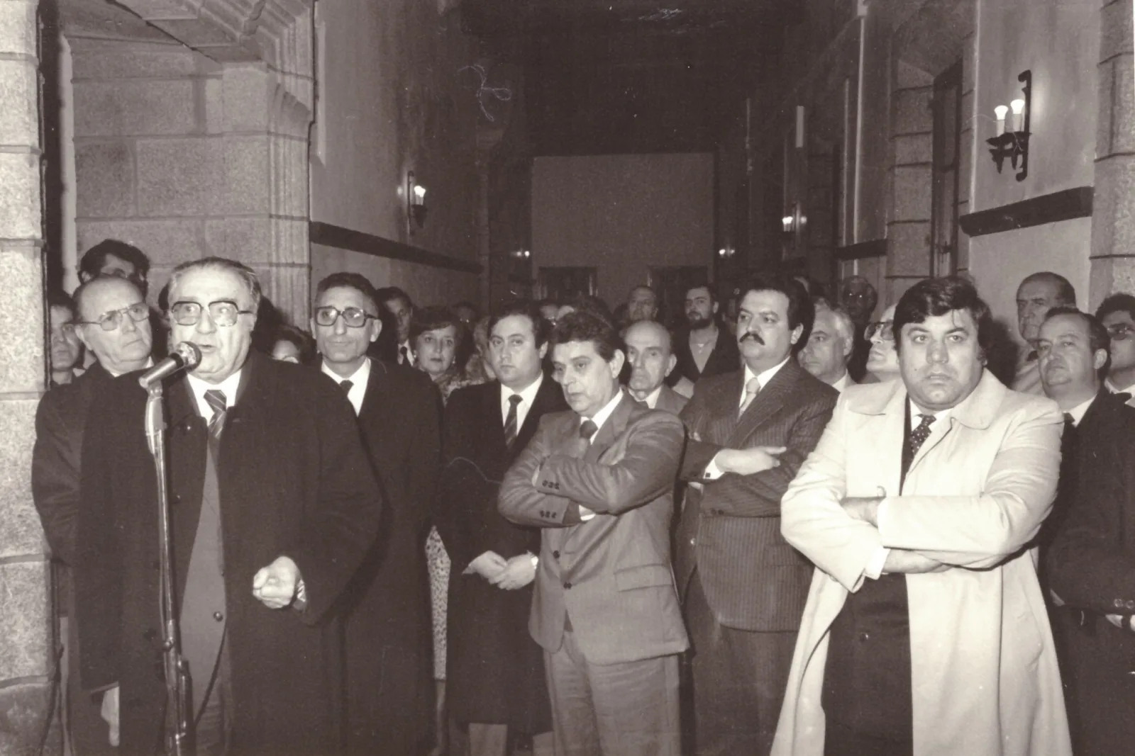 Fotografía en blanco y negro de un numeroso grupo de personas en un corredor de piedra escuchando a un hombre que habla ante un micrófono