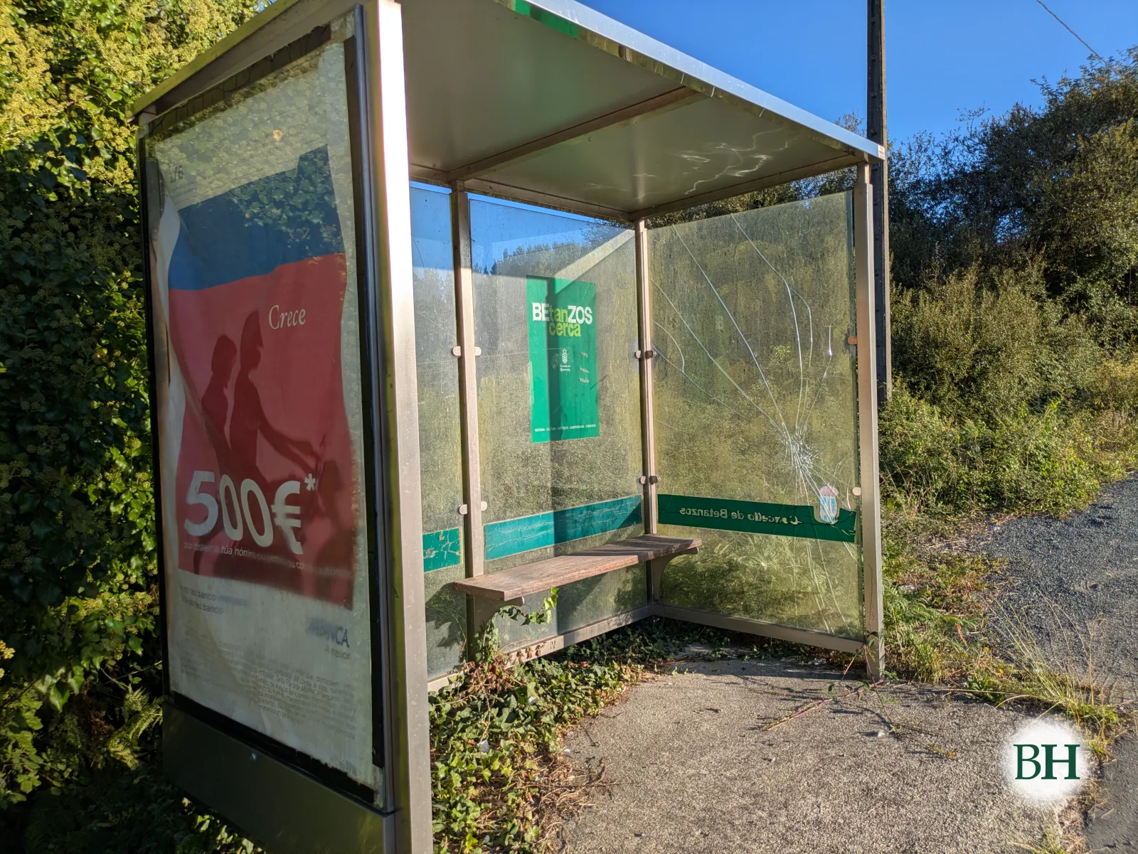 Marquesina de parada de autobús con un panel lateral de vidrio roto, banco de madera y cartel publicitario descolorido, rodeada de vegetación.