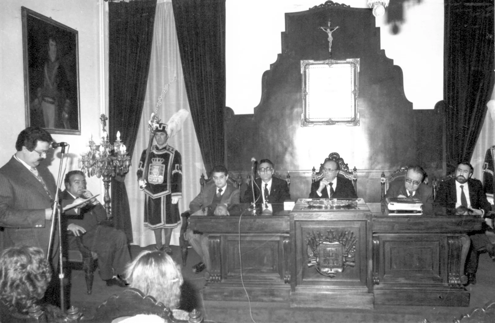 Fotografía en blanco y negro de un acto en la Sala Capitular del Ayuntamiento de Betanzos: un hombre lee ante un micrófono mientras las autoridades ocupan la mesa presidencial