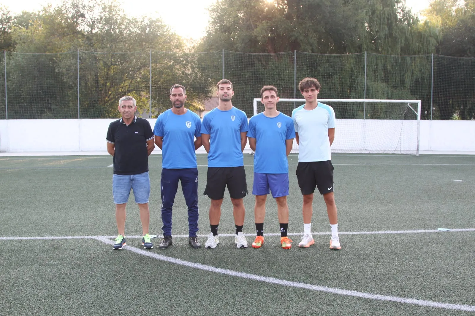 Cinco hombres del cuerpo técnico del Betanzos CF posan en fila sobre el césped artificial, delante de una portería, al atardecer