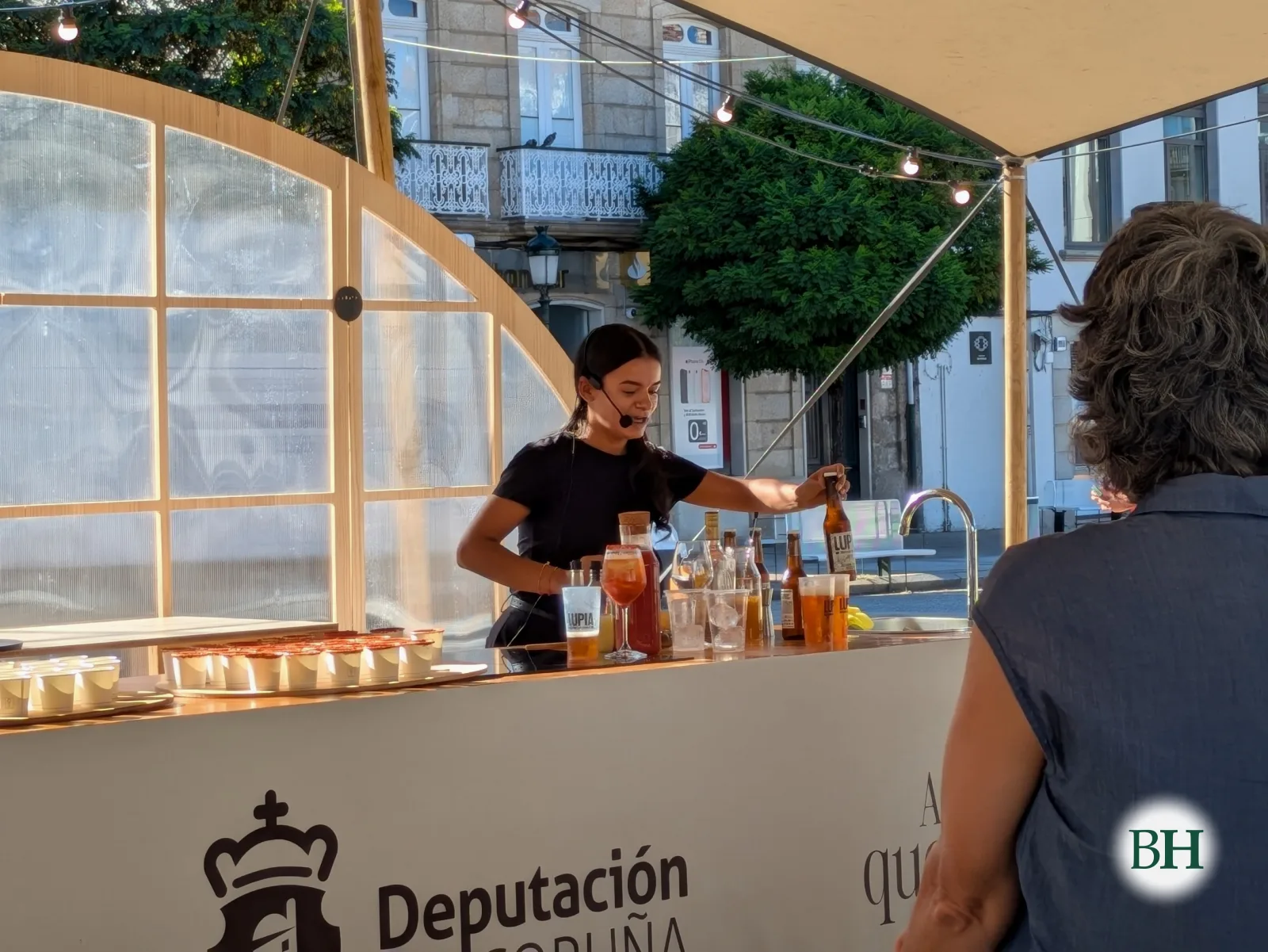 Una cocinera sirve una copa en un showcooking en la barra de la carpa de la Diputación de A Coruña, con botellas de Lupia delante