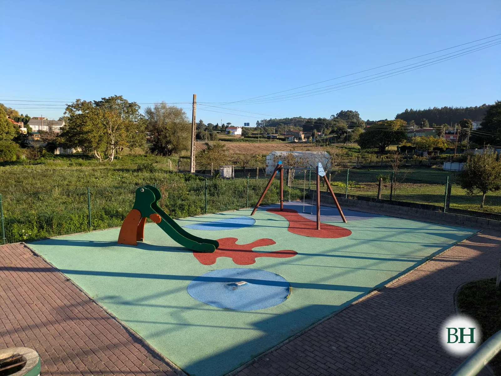 Parque infantil vallado con tobogán y columpio sobre pavimento de caucho de colores, con fincas y viviendas al fondo.