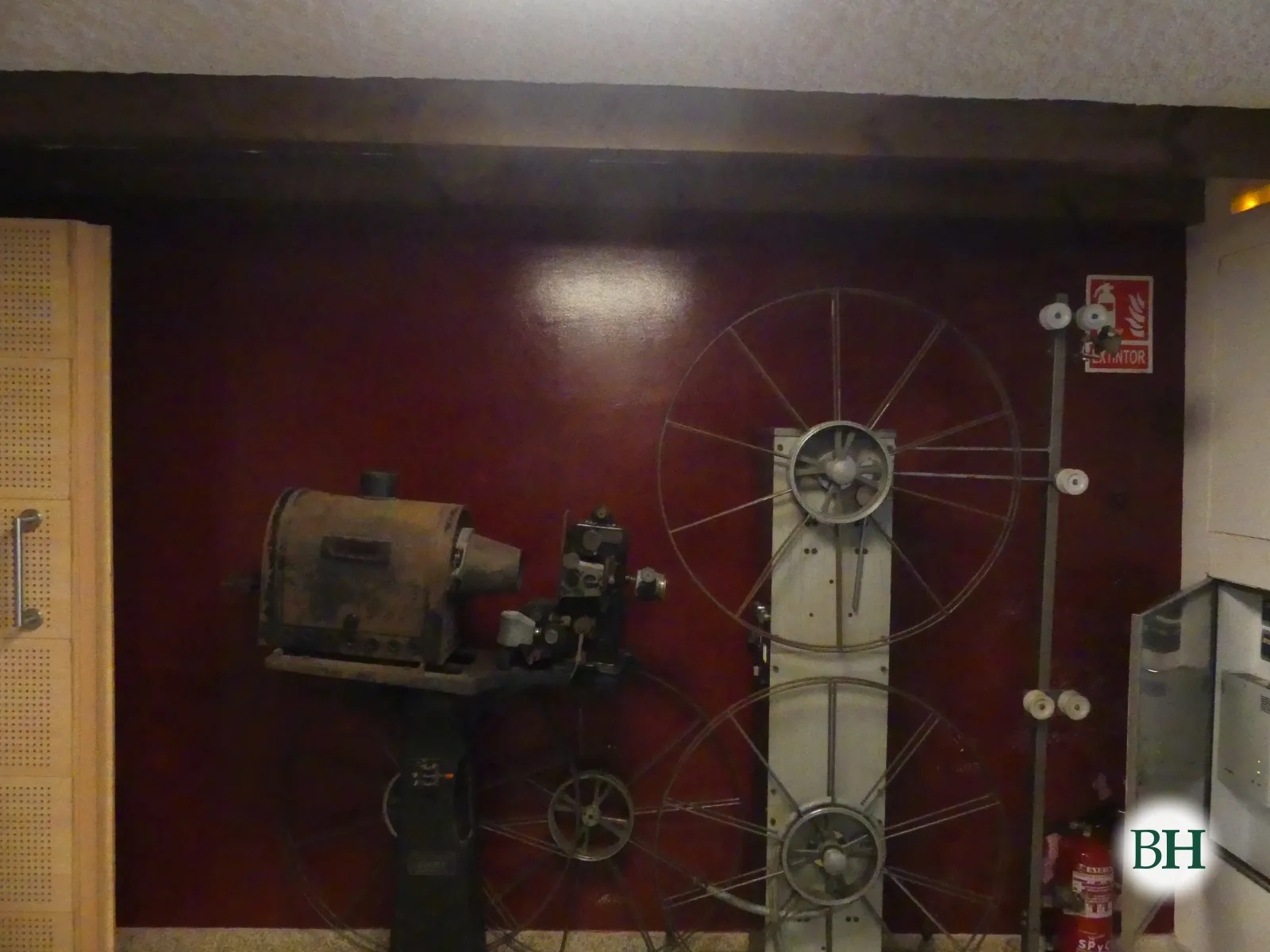 Un proyector cinematográfico antiguo y dos grandes bobinas expuestos contra una pared granate.