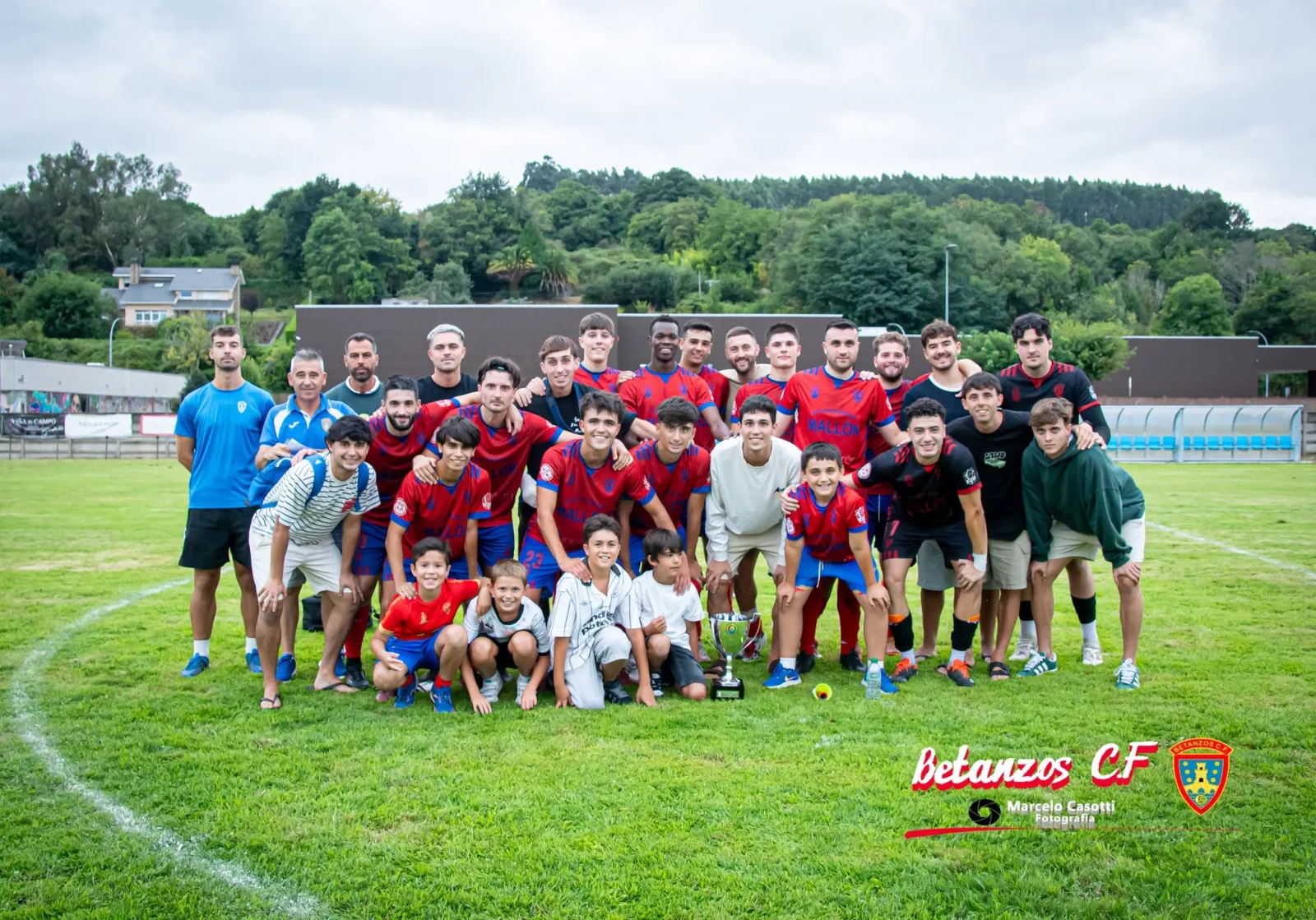El primer equipo del Betanzos CF posa sobre el césped con un trofeo en el suelo, rodeado de varios niños