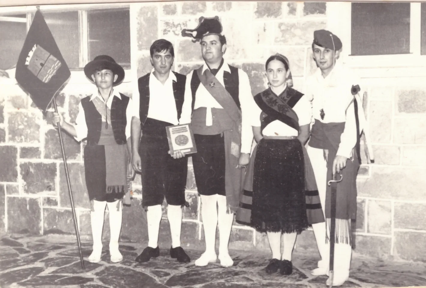 Fotografía en blanco y negro de cinco integrantes de un grupo de danza con traje tradicional, con una bandera y una placa, ante un muro de piedra