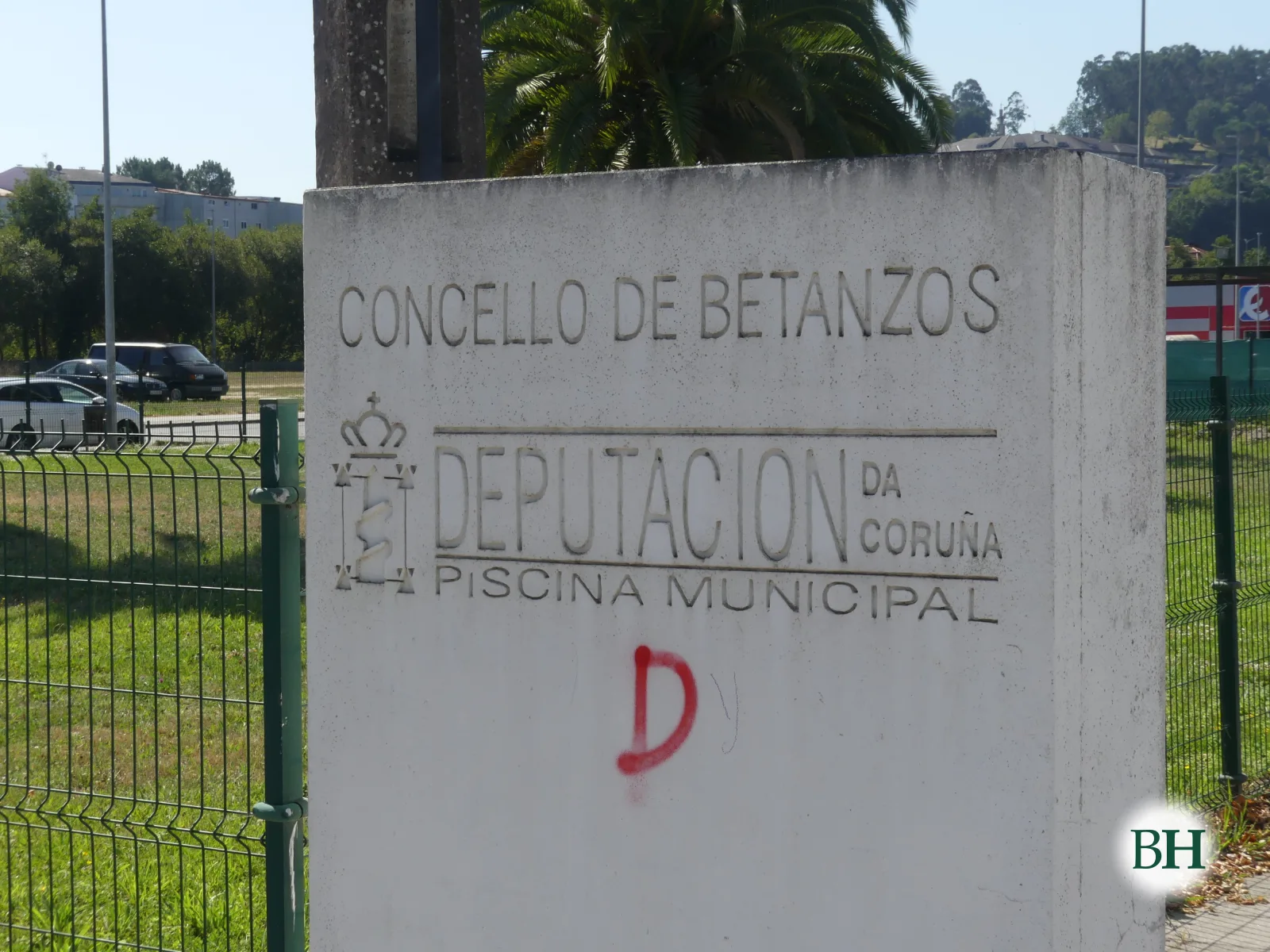 Monolito de piedra grabado con el texto CONCELLO DE BETANZOS, DEPUTACIÓN DA CORUÑA, PISCINA MUNICIPAL, junto a valla verde y palmera.