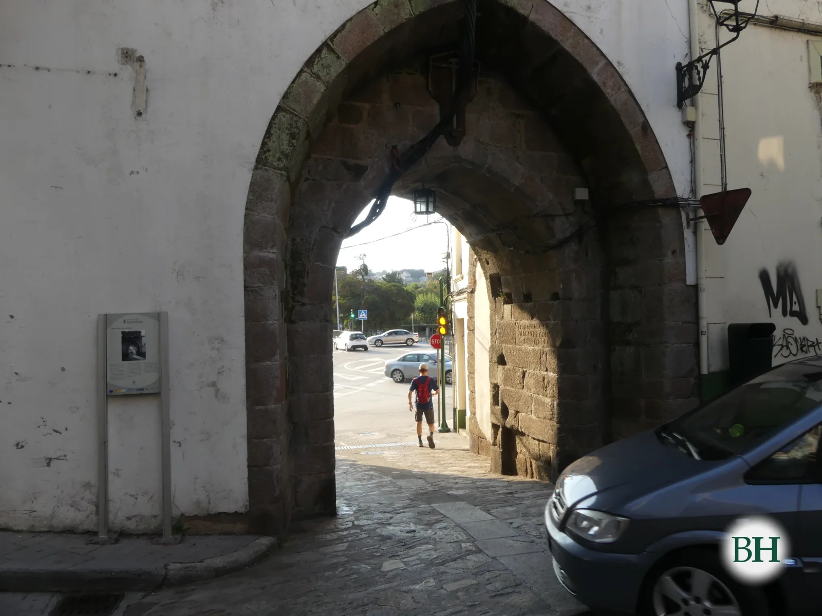 Vista más cercana del mismo arco de piedra, con un peatón con mochila roja al fondo y un coche aparcado en primer plano.