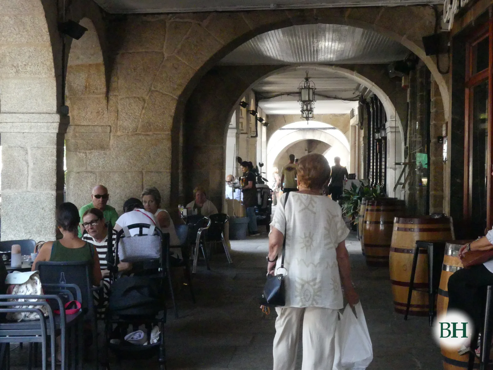 Galería de piedra con sucesión de arcos, terraza de bar con mesas, sillas y barriles de madera, y varias personas sentadas o caminando.