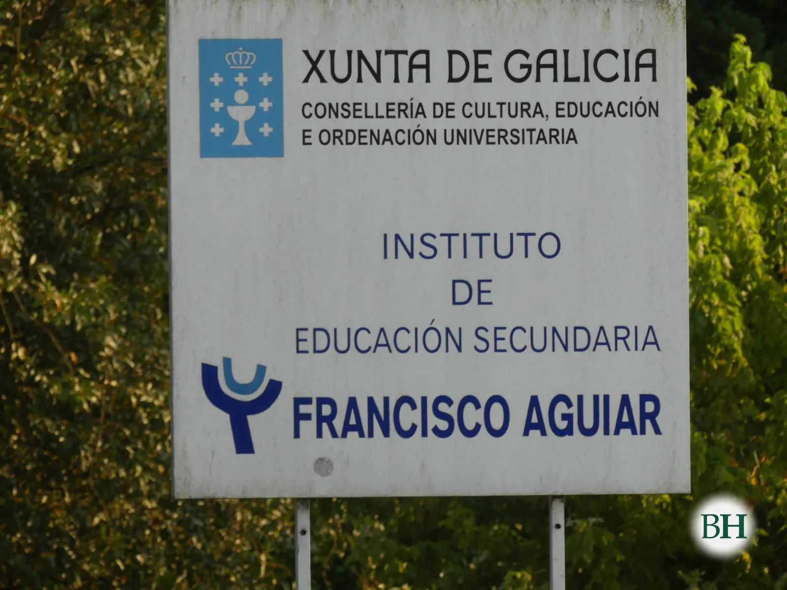 Cartel institucional branco con escudo e o texto Xunta de Galicia, Consellería de Cultura, Educación e Ordenación Universitaria, Instituto de Educación Secundaria Francisco Aguiar, entre vexetación.