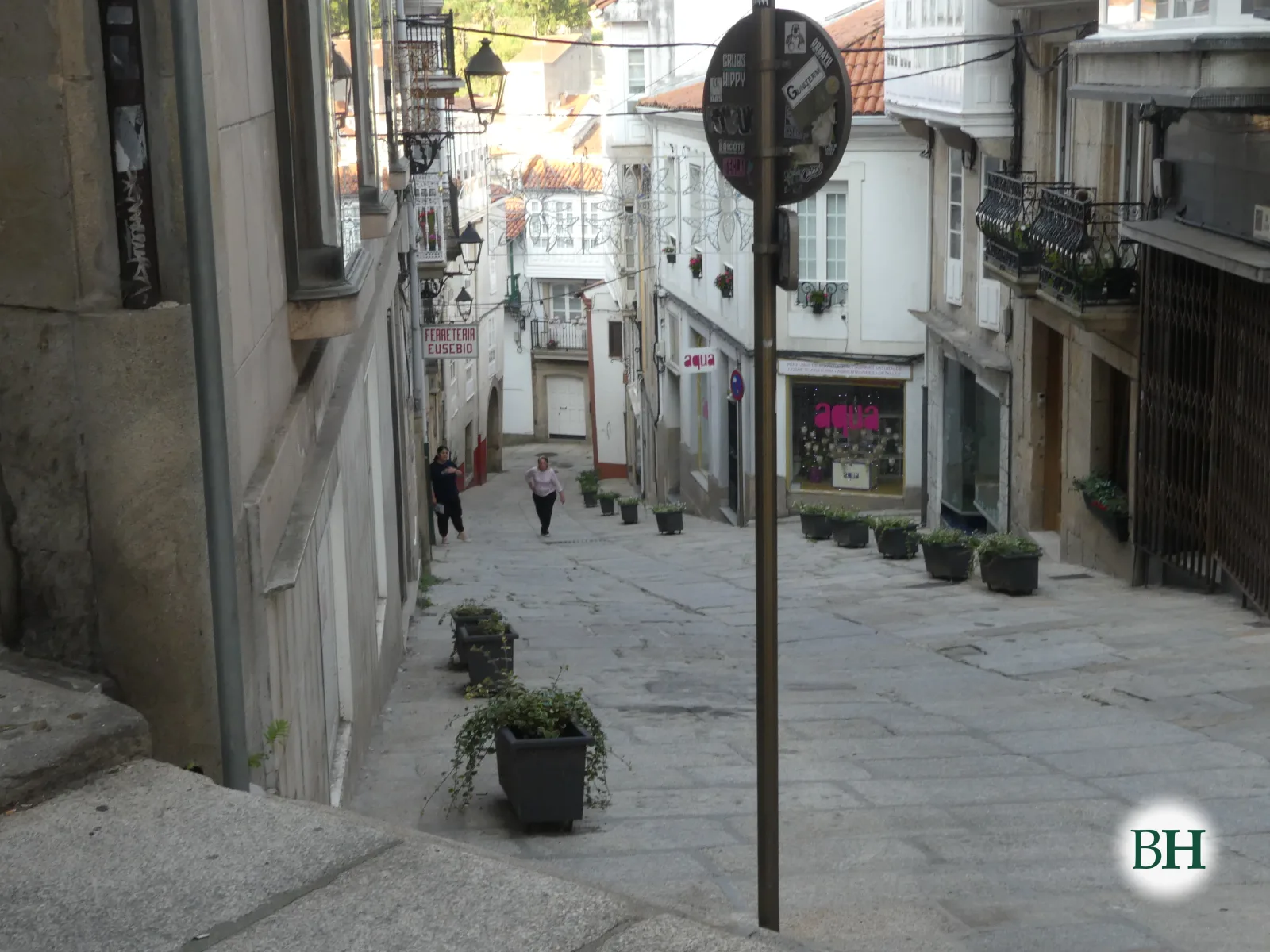 Rúa peonil en pendente con macetas de plantas, farola metálica e o rótulo dunha ferretería, con dúas persoas camiñando.