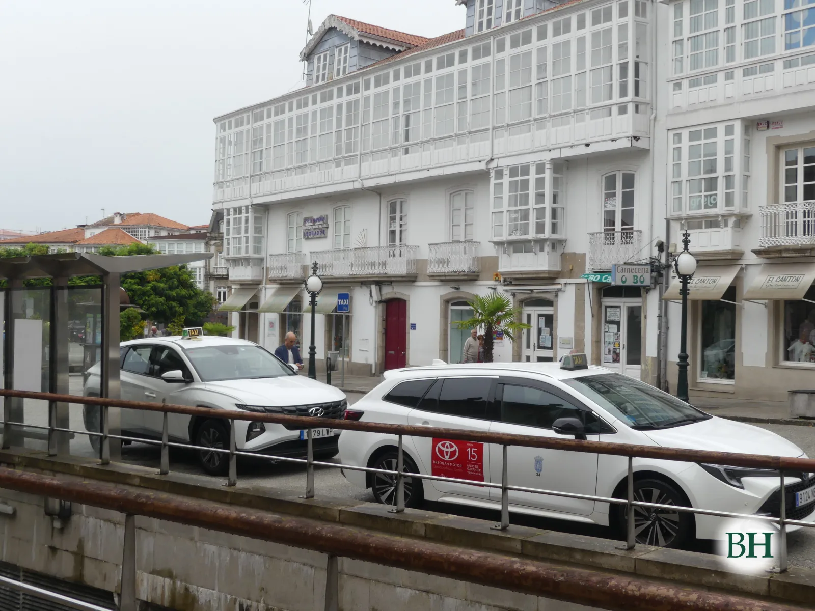 Parada de taxis con dous coches brancos fronte a un edificio de galerías acristaladas e unha palmeira.