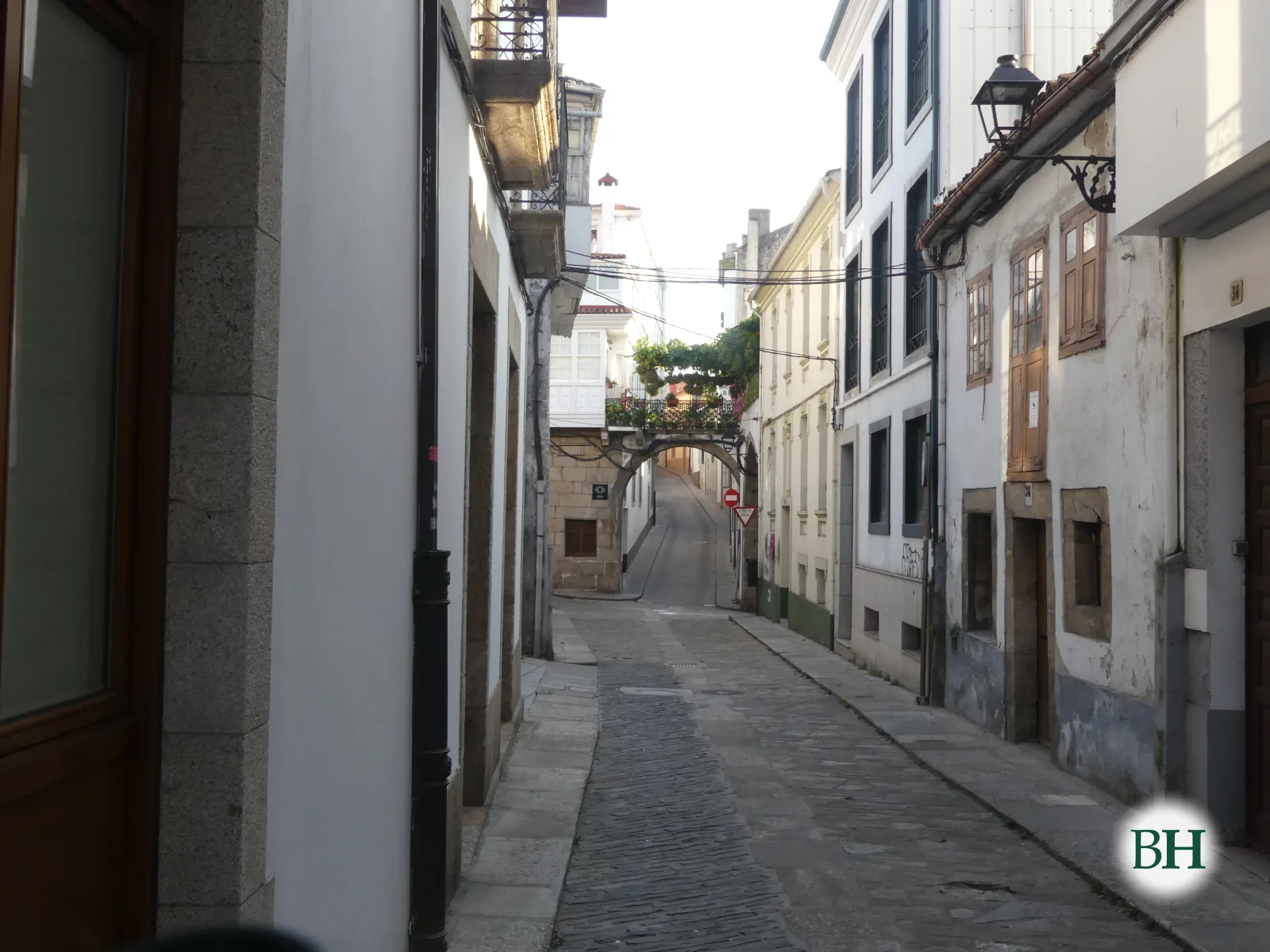Rúa estreita empedrada entre edificios con balcóns de ferro, farol de parede e un arco decorado con plantas ao fondo.