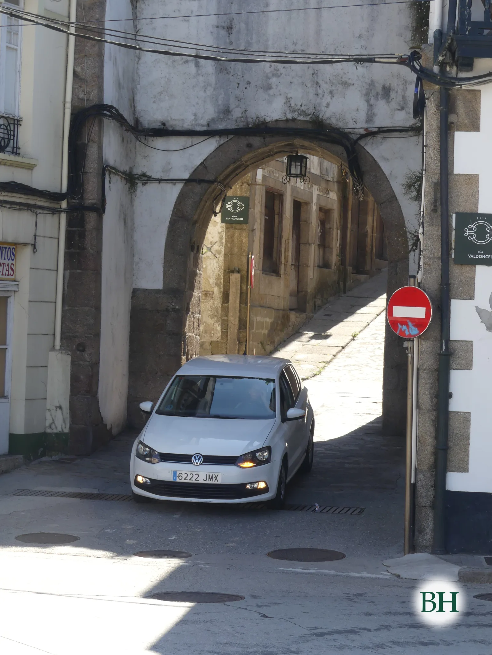 Coche branco circulando baixo un arco de pedra cun sinal de dirección prohibida, nunha rúa estreita de edificios encalados.