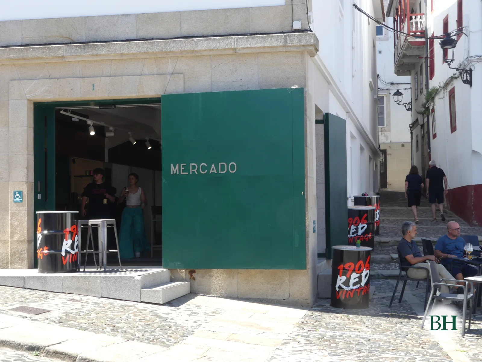Fachada de edificio con panel verde e rótulo MERCADO, rúa empedrada con farolas, terraza con mesas e persoas sentadas.