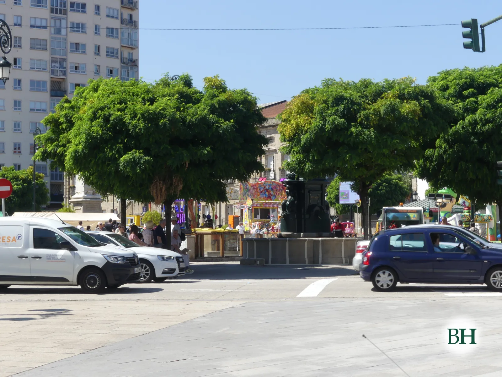 Praza urbana con edificio de apartamentos ao fondo, árbores frondosas, fonte central, postos de feira e coches circulando baixo ceo despexado.