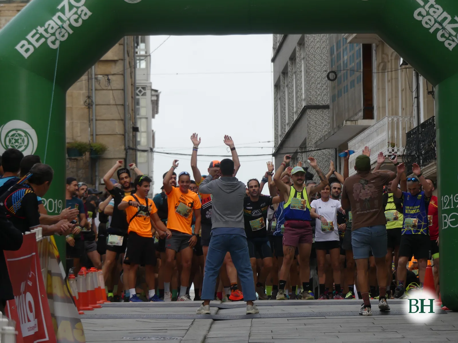 Corredores de pé cos brazos en alto celebrando baixo o arco de saída mentres o speaker e un fotógrafo os dirixen.