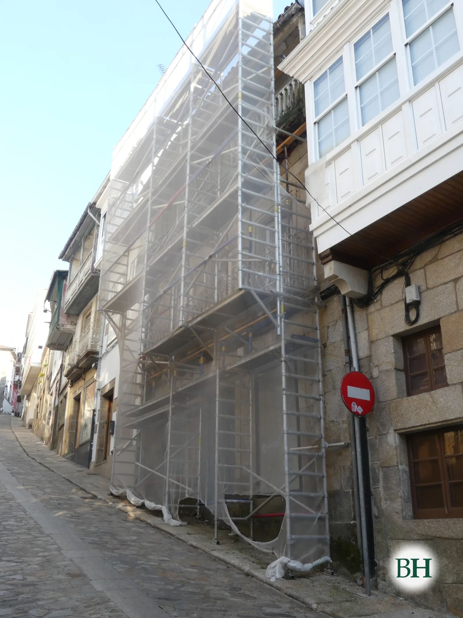 Fachada en rehabilitación cubierta por andamio metálico con lona translúcida, junto a una casa con galerías acristaladas blancas, en calle empedrada en cuesta.