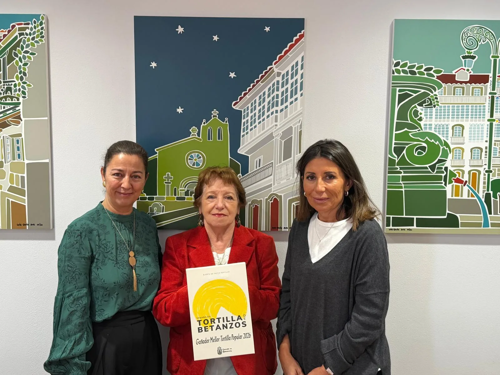 Tres mujeres posan ante unos cuadros con vistas de Betanzos. La del centro sostiene el diploma de ganador de la Mejor Tortilla Popular 2026 de la Semana da Tortilla de Betanzos.