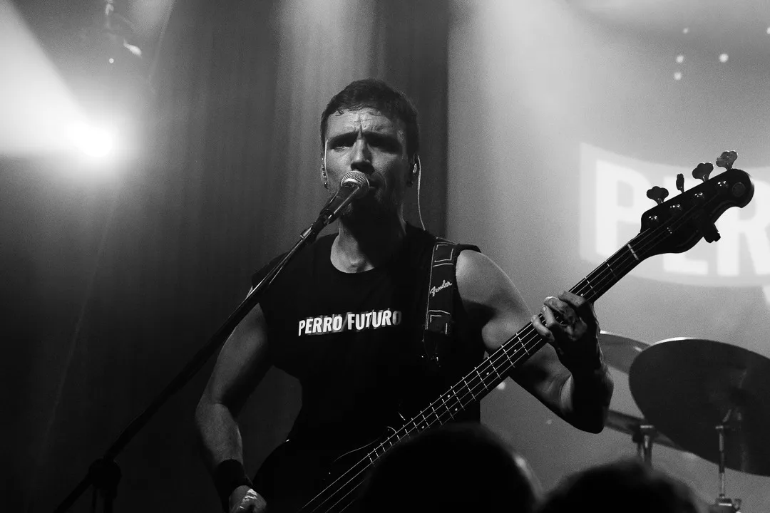 Diego Amigo, bajo y voz de Perro Futuro, canta ante el micrófono con una camiseta con el nombre del grupo, en una foto de concierto en blanco y negro.