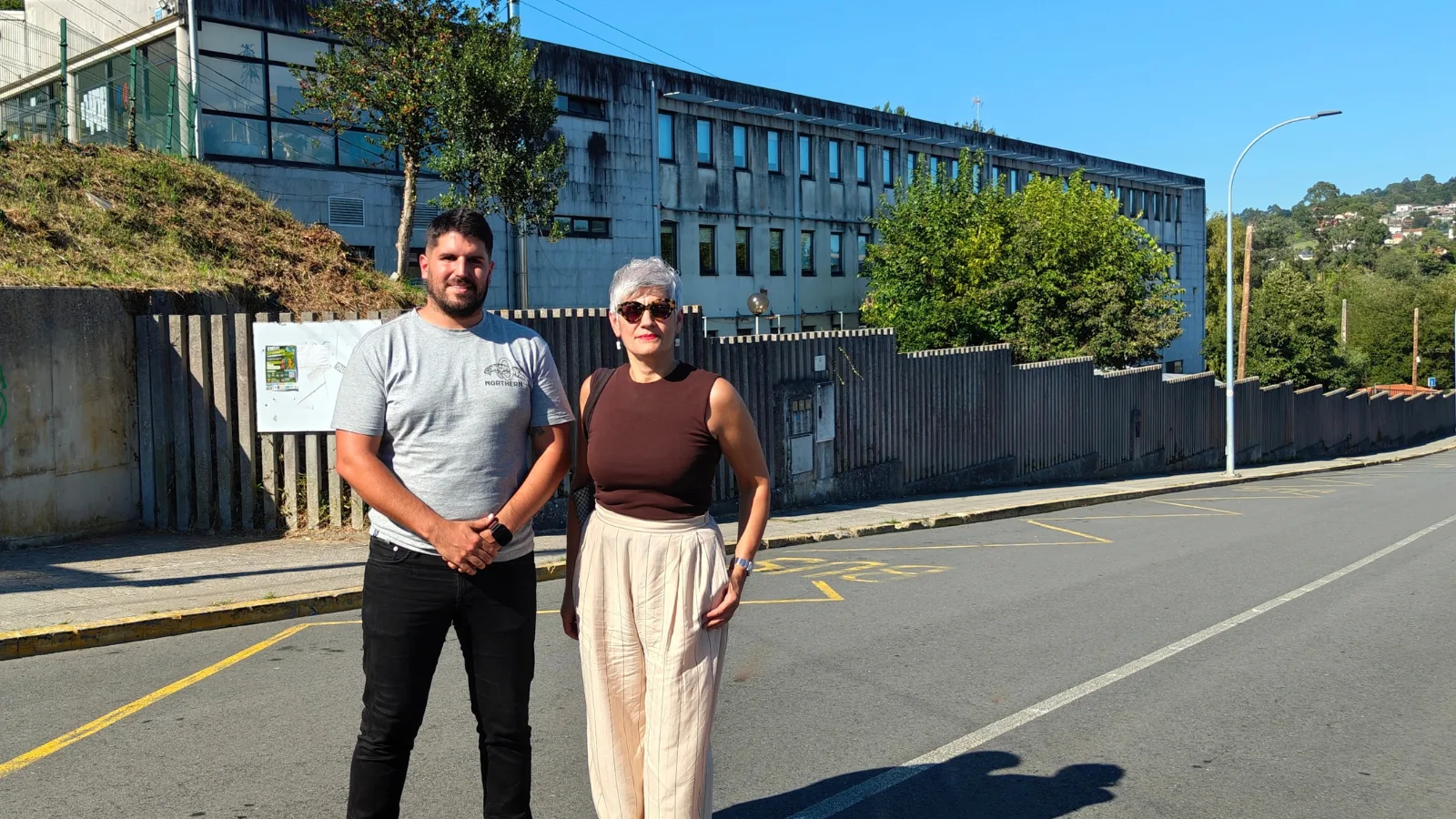 Alberte G. Nicolás, con una camiseta gris, y Mercedes Queixas, con gafas de sol, de pie en una calle delante del edificio del CPI Castro Baxoi, en Miño.