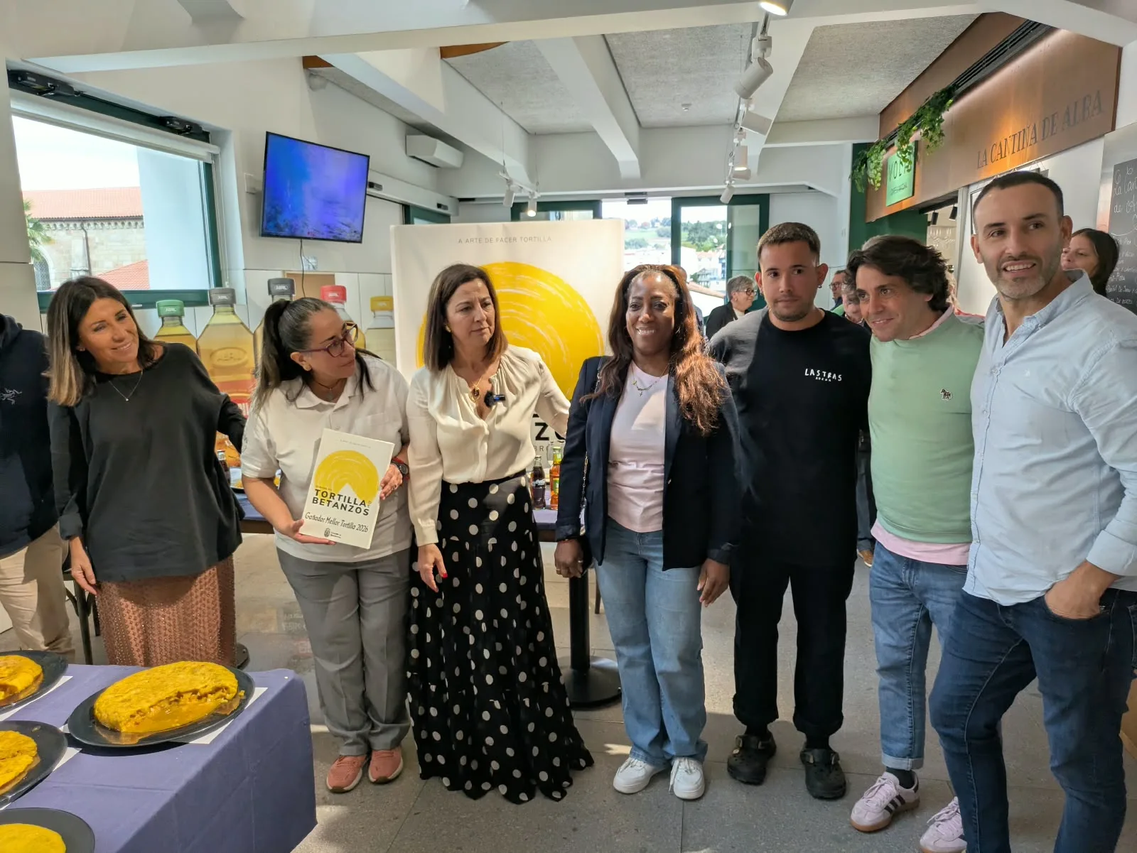 Nueve personas posan de pie y sonrientes en un local de paredes blancas y techo alto, delante de un cartel amarillo de la Semana de la Tortilla. Una mujer del centro sostiene un diploma de la Tortilla de Betanzos. Delante, una mesa con mantel azul y