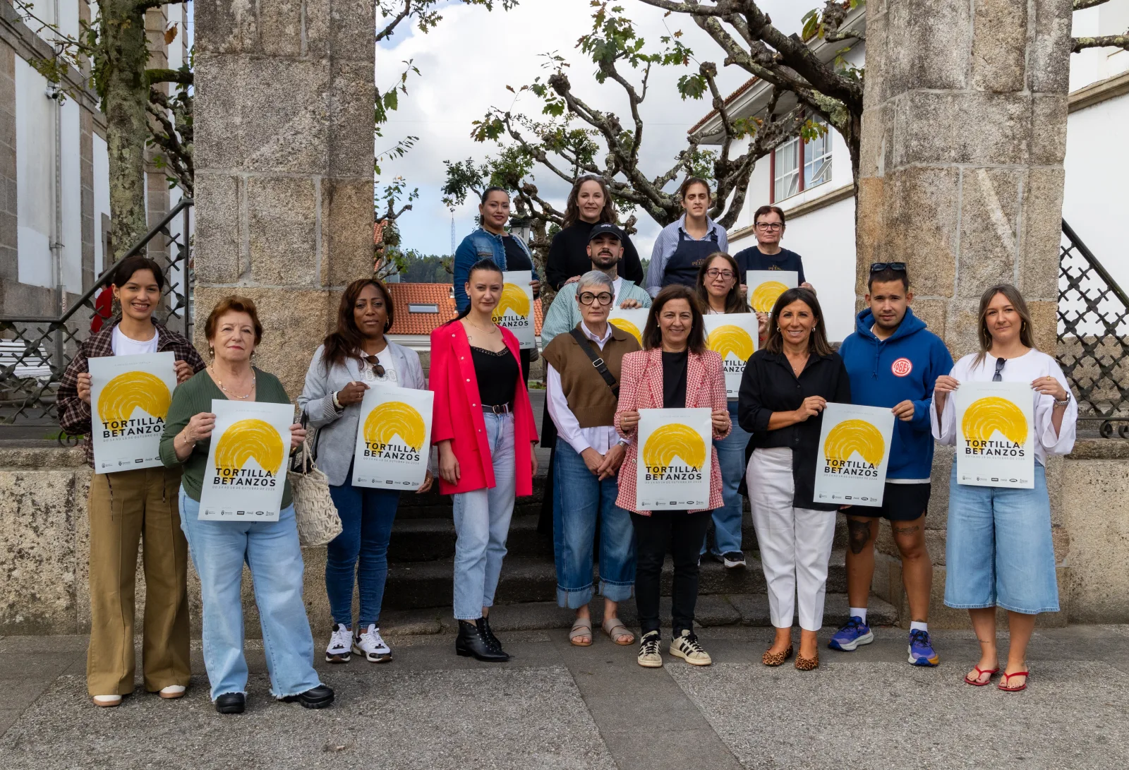 Presentación de la XVI Semana de la Tortilla de Betanzos: una quincena de personas, hosteleras y hosteleros participantes y representantes municipales, posan entre dos pilares de piedra sosteniendo el cartel amarillo del evento.