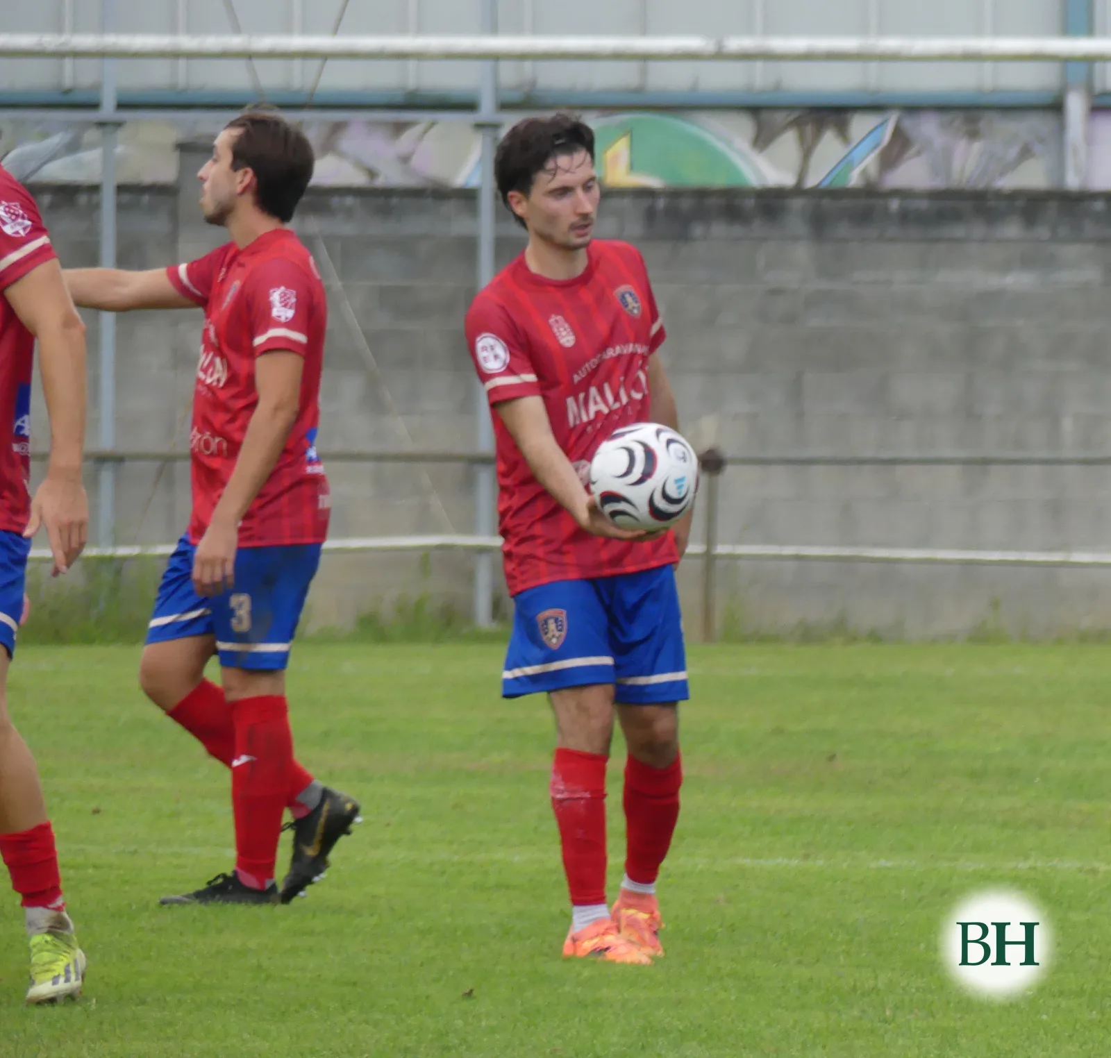 Tres jugadores del Betanzos CF, de camiseta roja y pantalón azul, en el césped. El del centro sostiene el balón con una mano.