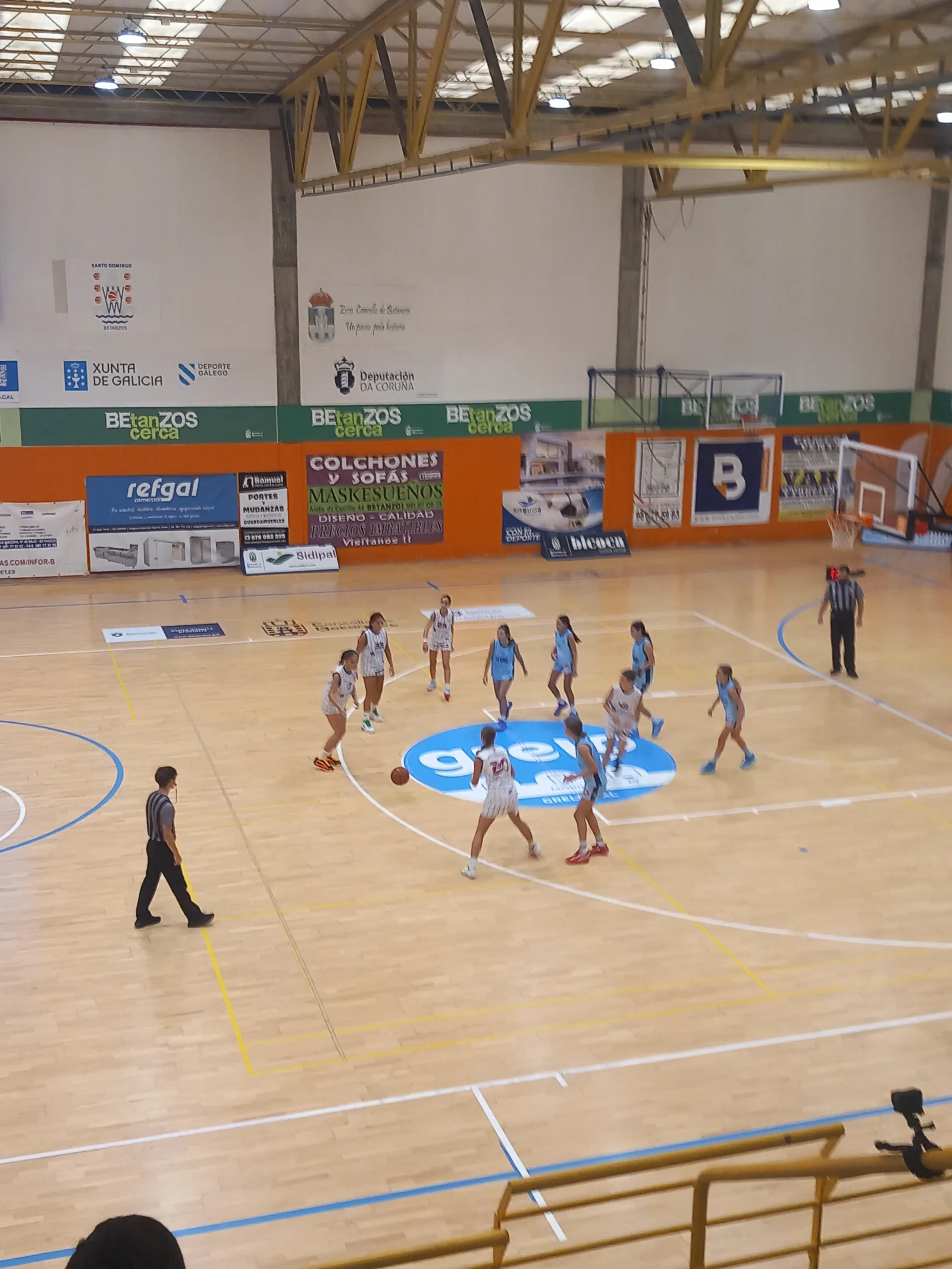 Una jugadora del infantil femenino del Santo Domingo, de blanco, bota el balón en el círculo central del pabellón de Betanzos ante el Estudiantes Lugo, de celeste.