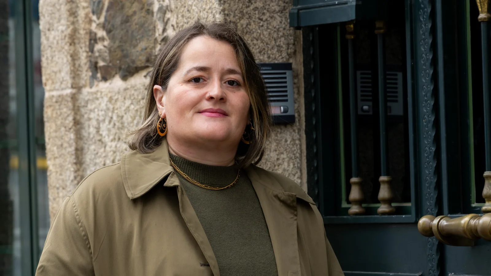 La portavoz municipal del BNG de Oza-Cesuras, Estefanía Busto, con una gabardina verde oliva, delante de una pared de piedra y una puerta de hierro.