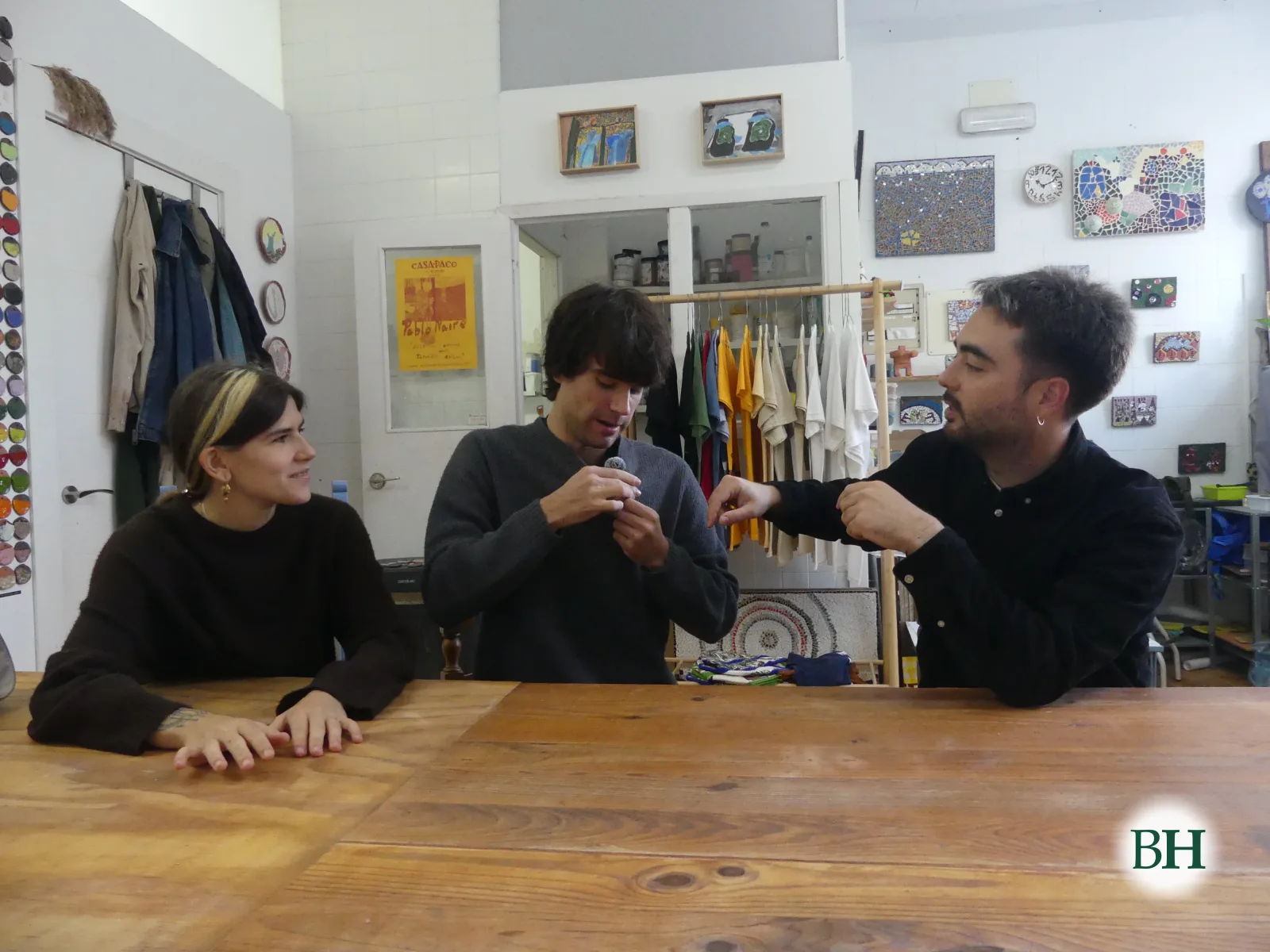 Andrea Aguafría y los hermanos Pablo Naire y Miguel, sentados a una mesa de madera en el estudio Río Río, en Betanzos. Al fondo, un burro con camisetas de colores, un cartel amarillo y mosaicos pequeños en la pared.