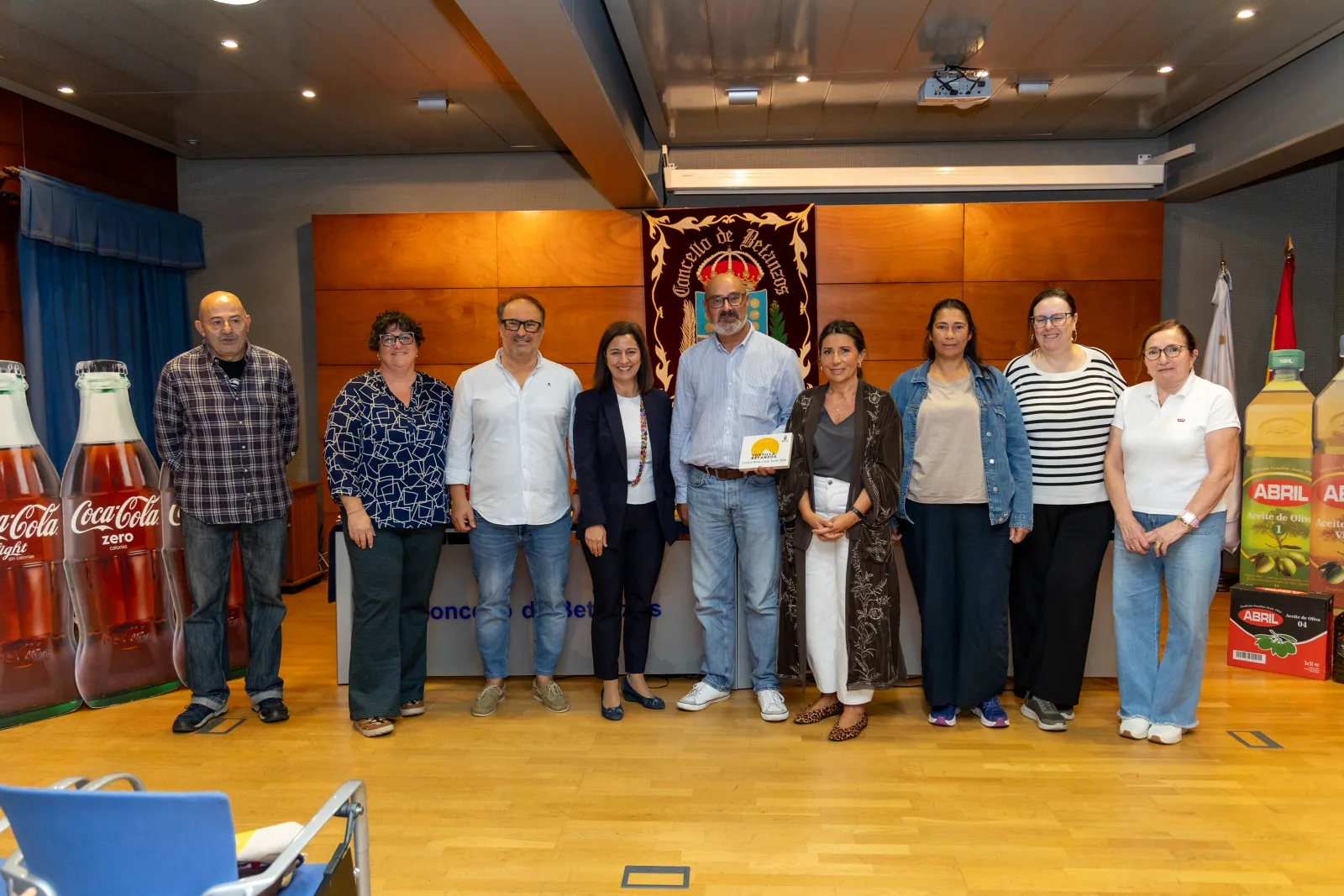 Foto de familia de once personas de pie en un salón con pared de madera, delante de un repostero del Concello de Betanzos. Un hombre del centro tiene un diploma. A los lados, botellas gigantes de refresco y de aceite.