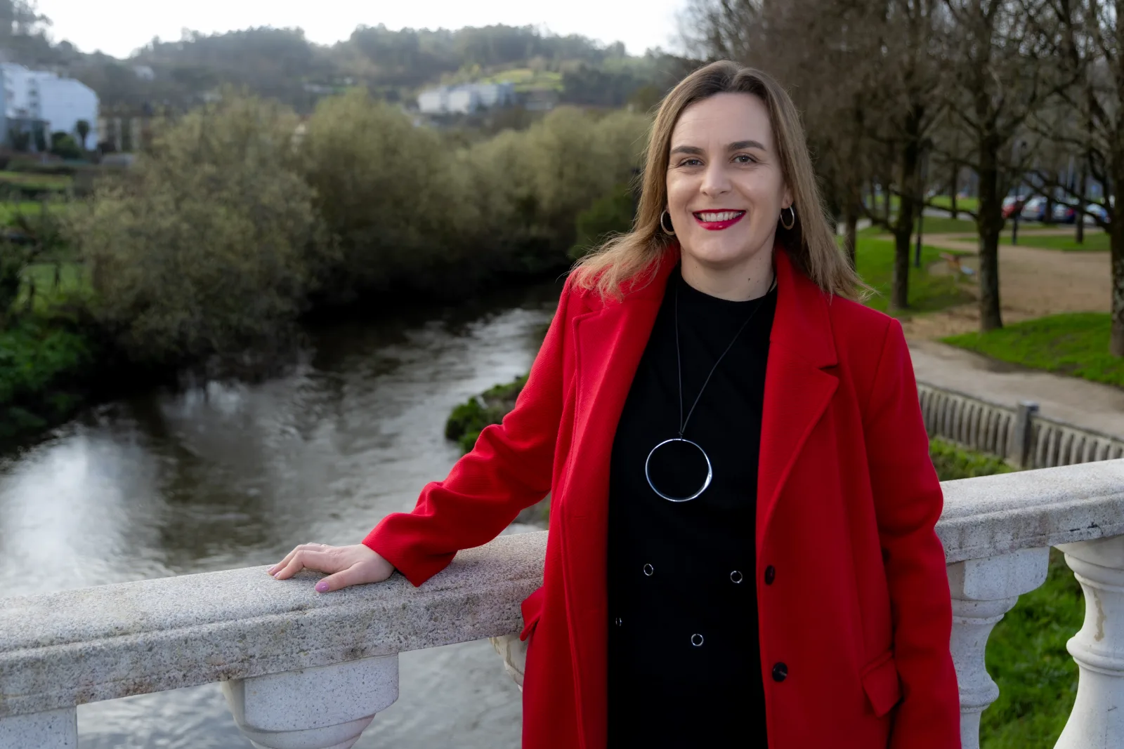 A concelleira do BNG de Betanzos Amelia Sánchez, cun abrigo vermello, apoiada no varandal dunha ponte sobre un río.