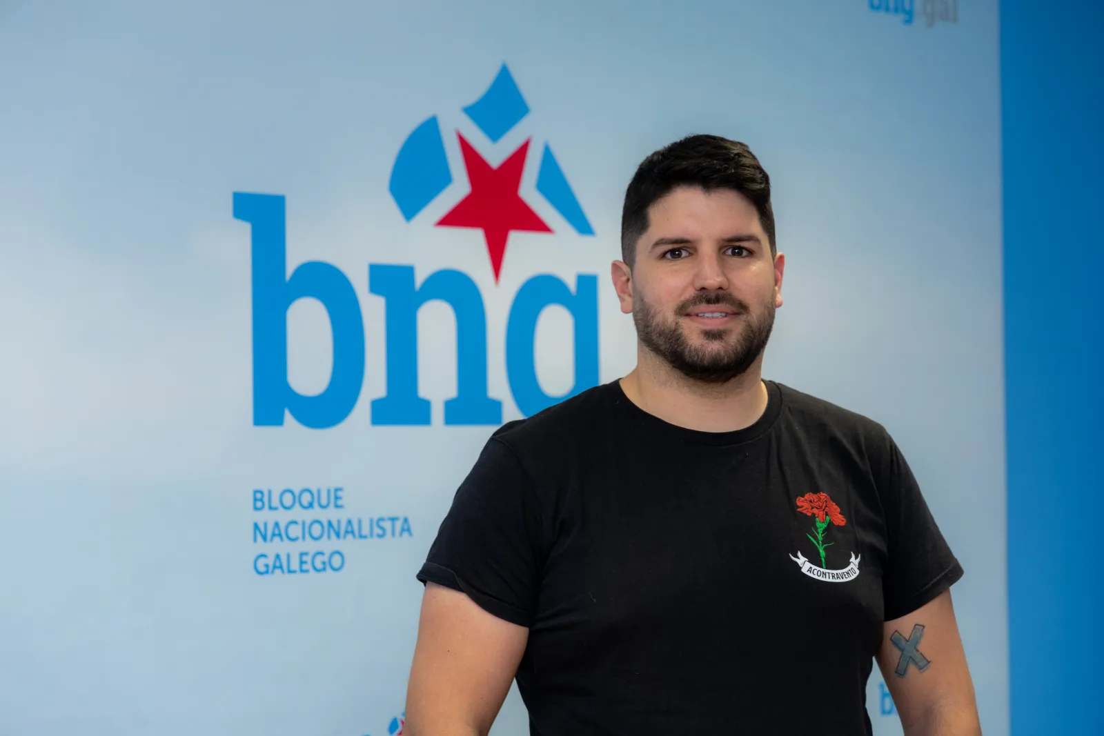 O portavoz municipal do BNG de Miño, Alberte G. Nicolás, cunha camiseta negra, diante dun panel azul co logotipo do BNG.